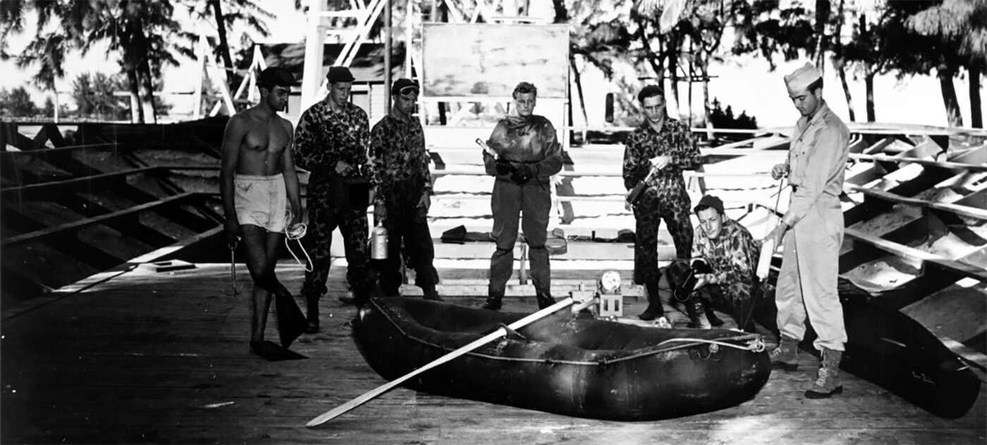 Personnel demonstrate the inflation of a small rubber boat, 10 December 1943. Other men stand by with a variety of equipment. Note: camouflage uniforms; swimming fins and mask on one man, who holds a grappling hooks; rubber diving suit on man in center; under water lights; and kayak "your baby" at far right.