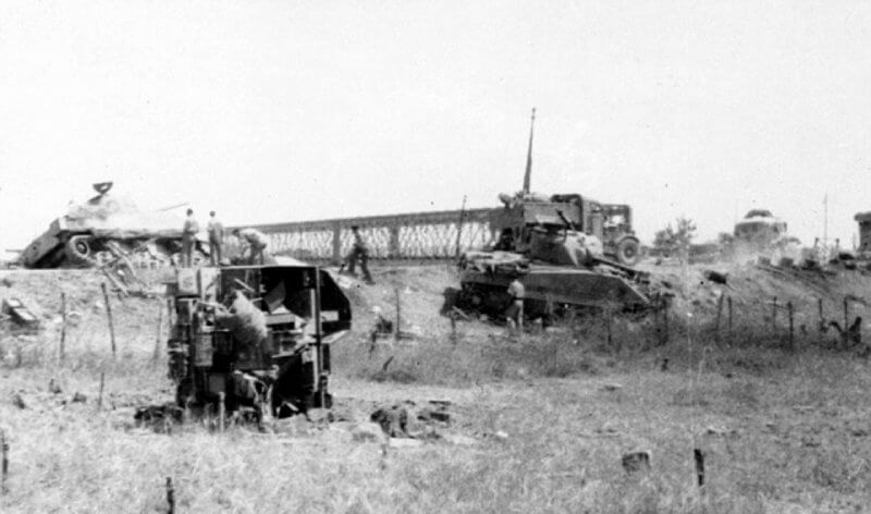 The view from the southern bank of the Primosole Bridge, captured after the battle, highlights the aftermath of fierce combat. The embankment of the highway, prominent in the photograph, posed tactical challenges for troops during the engagement—difficult terrain to navigate and fortify, both in reality and within historical interpretations.
Two Sherman tanks are visible on the northern bank, likely casualties of accurate fire from 88mm guns defending the Axis positions. To the right in the background, one of the pillboxes guarding the bridge can be seen, a reminder of the fortifications that made this location a focal point of resistance.
In the foreground, wire obstacles and a capsized carrier suggest the intensity of the fighting.