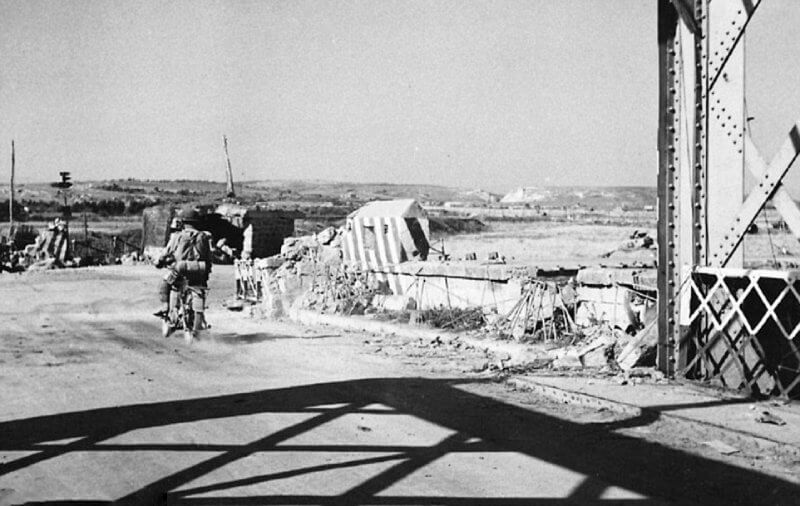 The view from the southern end of the Primosole Bridge reveals the "Johnny" hills in the background, a critical high ground that played a strategic role in the battle. Visible in the scene is one of the two pillboxes that had been part of the Axis defensive line, constructed to oversee and defend this vital crossing point.