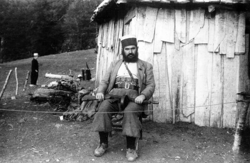 SERVICE OF MAJOR DAVID SMILEY WITH SPECIAL OPERATIONS EXECUTIVE (SOE) IN ALBANIA, 1943 -    1944. (HU 65033) First mission: local 'ceta' leader Baba Faja (with Thompson sub-machine gun) seated    outside the wooden HQ building at Bixha, October 1943. Copyright: ý IWM. Original Source: http://www.iwm.org.uk/collections/item/object/205061235