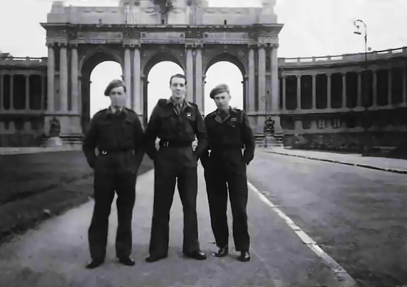 Captain Johnny Cooper (centre) and Sergeant Jeff Du Vivier (right) stand in front of the Arch of the Cinquantenaire in Brussels, captured in a moment of quiet before the final push into Germany. It is early 1945, and the men, veterans of months of behind-the-lines warfare, are part of a newly formed composite force drawn from 1 and 2 Special Air Service Regiments.