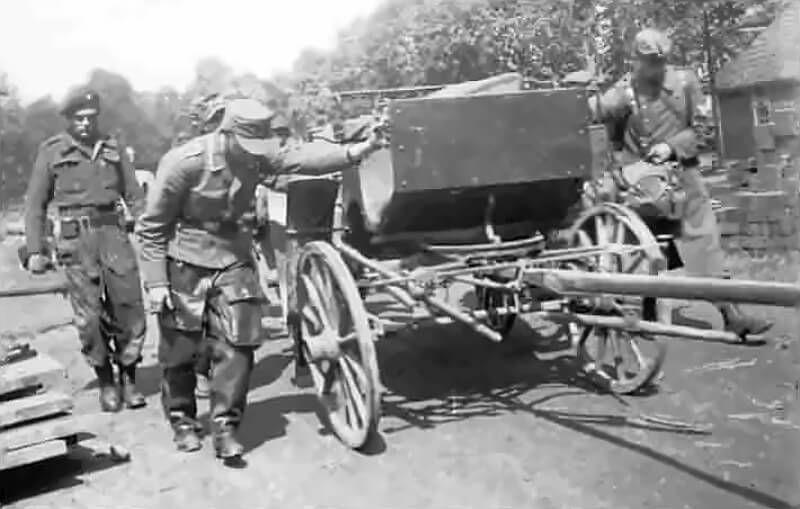Lieutenant Denis Wainman (left) of C Troop, 1 Special Air Service stands at the roadside, watching with a measured gaze as a group of captured German soldiers push a wooden carriage through a quiet, war-weary village in rural Germany.