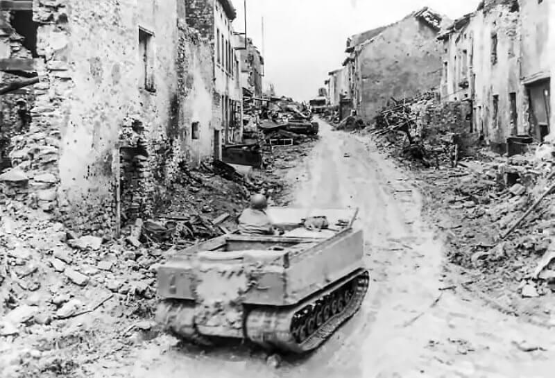 A basic M29A Weasel advances cautiously into a shattered village, rubble and destroyed buildings lining the street.